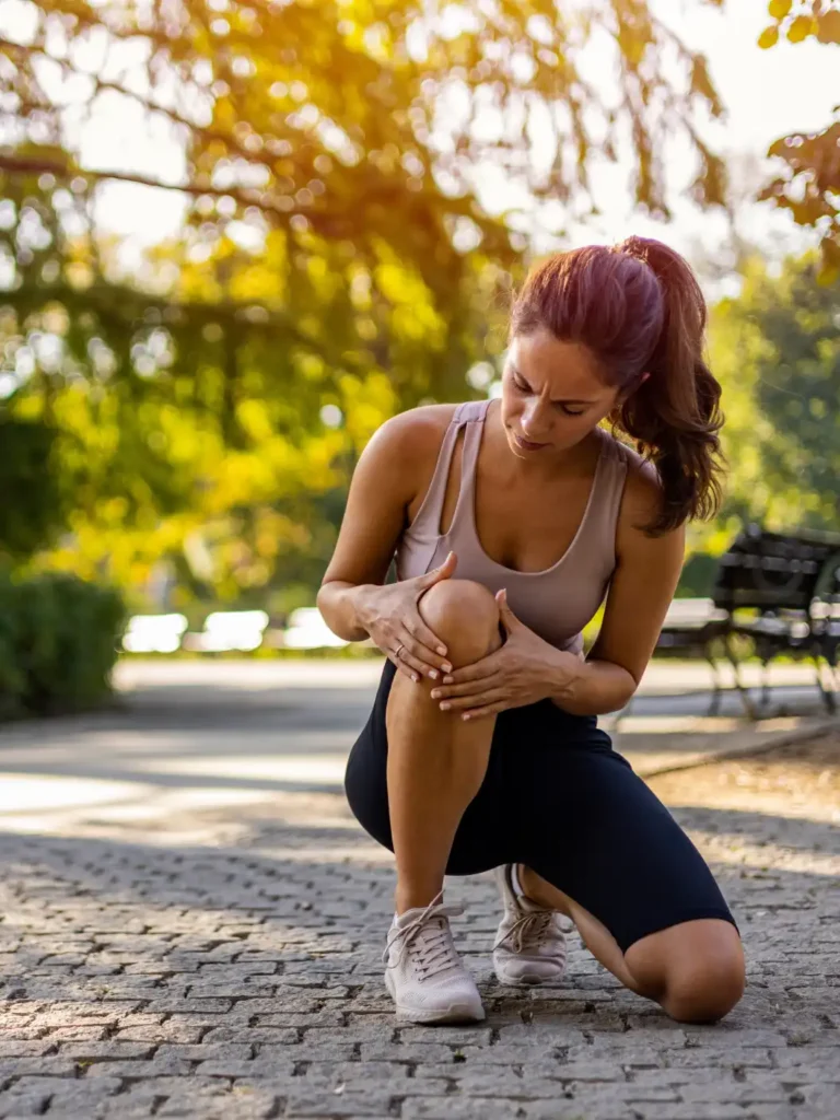 A woman in workout clothes kneels on a paved path in a sunlit park, clutching her right knee with both hands in visible pain.